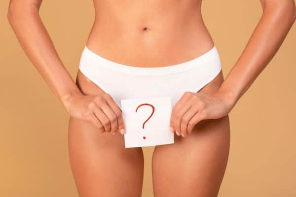 Mulher segurando um cartaz branco, no qual consta um ponto de interrogação, frente ao aparelho genital. Conceito de cirurgia íntima estética e funcional.