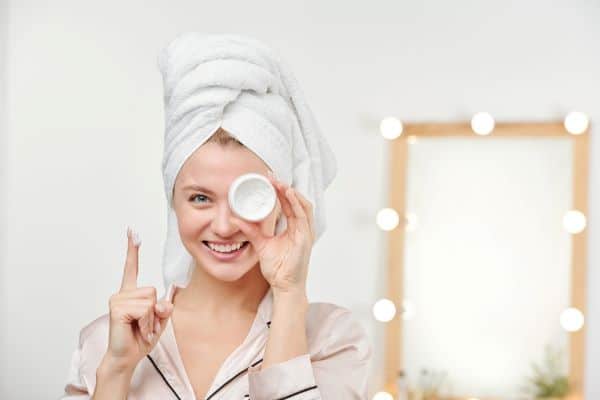 Jovem mulher, usando roupão e uma toalha enrolada na cabeça, segura um pote de creme facial frente ao olho esquerdo e aponta o dedo indicador da mão direita no qual se encontra uma porção de creme. Imagem faz alusão à rotina de cuidados com a pele da face, especialmente após procedimentos.
