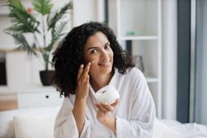 Bela mulher afro segurando um frasco de creme facial, aplicando-o no rosto.