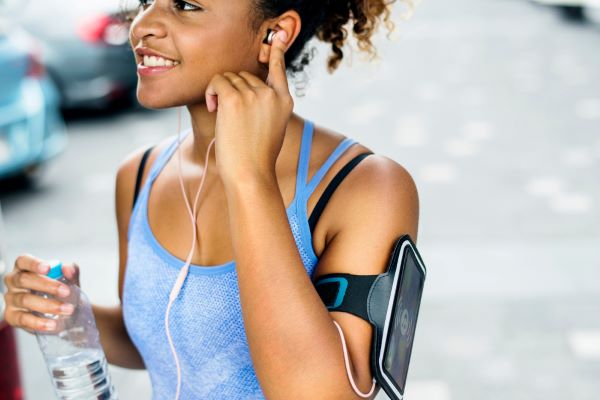 Mulher negra, jovem, praticando atividade física após 1 mês de procedimento estético. Para realizar a caminhada, ela carrega um a garrafa de água, seu celular e fones de ouvido.
