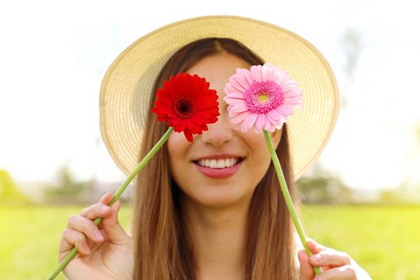 Jovem loira, com cabelos compridos, passeia pelo campo usando um chapéu e cobrindo os olhos com 2 gérberas (vermelha e rosa).
Conceito de cirurgias plásticas na primavera.