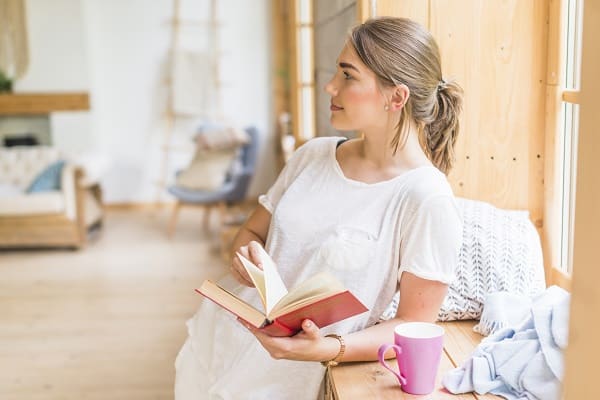 Mulher jovem, usando um vestido branco leve, encostada no peitoril de uma janela da sala, onde está uma caneca rosa, lê um livro.