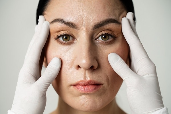 Mãos de um cirurgião plástico, usando luvas brancas, seguram o rosto de paciente para avaliar blefaroplastia e lifting de supercílios.