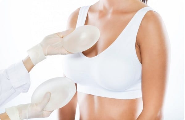 Médica usando luvas brancas avalia o volume ideal da prótese de silicone que será colocada em paciente. Ela segura 2 próteses frente ao colo da mesma.