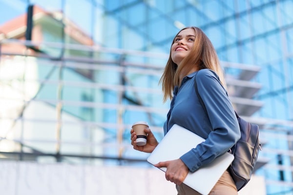 Um dos mitos do pós-operatório é a volta ao trabalho em 7 dias. Isso depende do procedimento. 
Na imagem, uma linda jovem, vestindo camisa azul , calça bege e mochila de couro, segura um copo de café e um notebook.